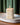 White Mexican sculpture candle with Talavera-inspired floral design burning on a white ceramic tray, set on a wooden surface in front of green curtains.