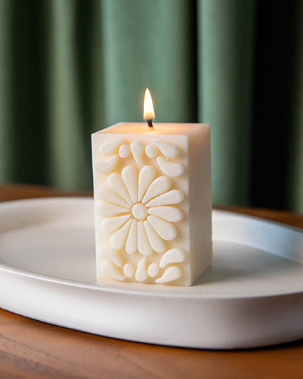 White Mexican sculpture candle with Talavera-inspired floral design burning on a white ceramic tray, set on a wooden surface in front of green curtains.