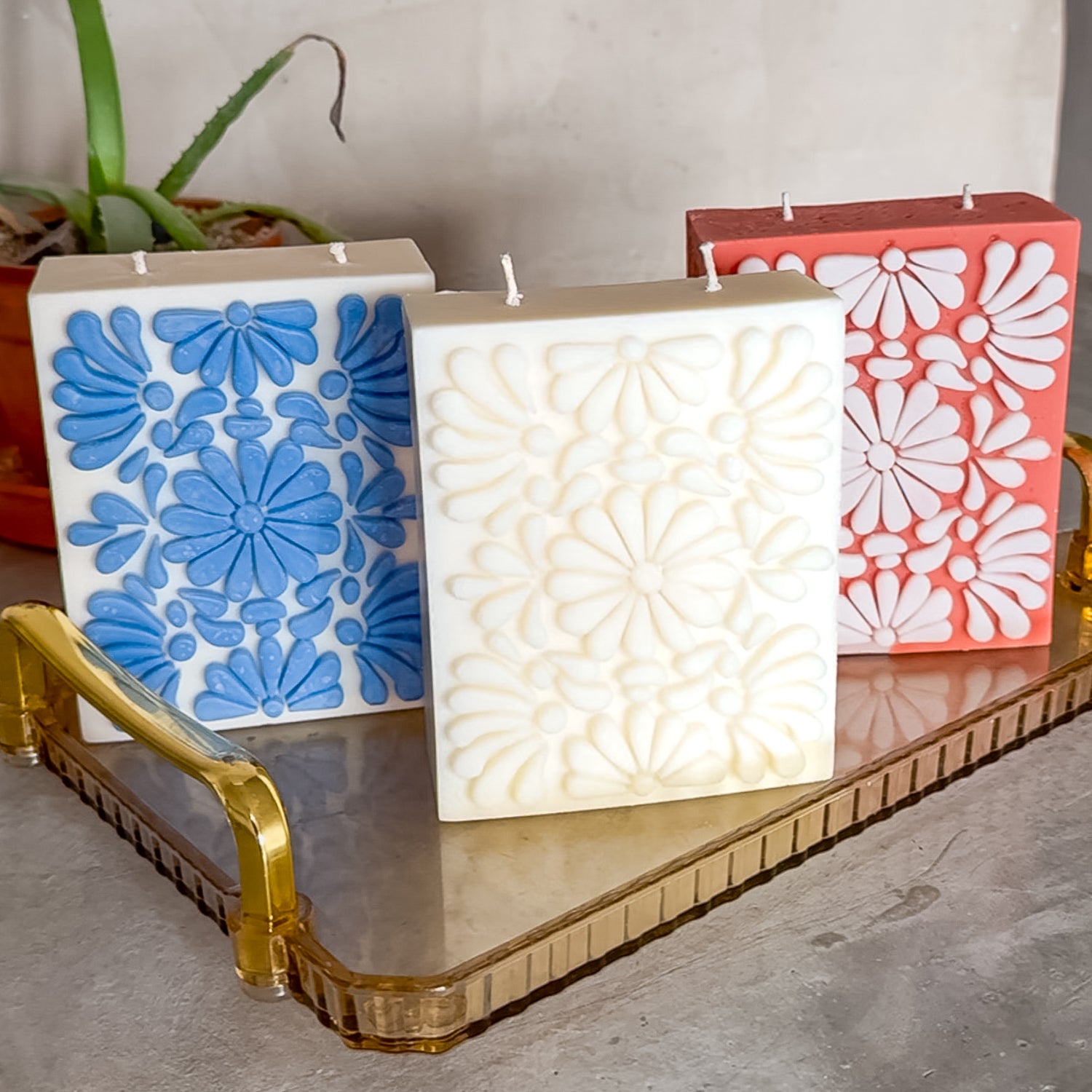 Trio of handcrafted Mexican candles featuring Talavera-inspired floral designs in blue, white, and terracotta, displayed on a vintage acrylic tray with gold handles, with a small potted plant in the background.